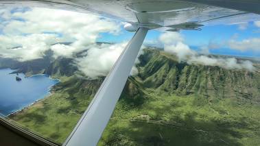 Small aircraft flies over Hawaii with aerial view of green, lush volcanic mountains.