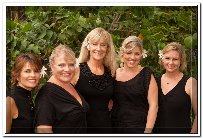 five women pose together for photo all wearing black in front of green plant background