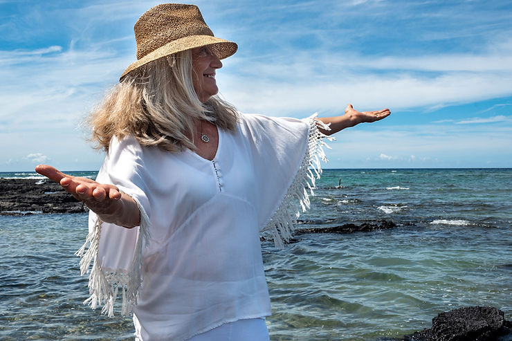 Woman in a white top and hat joyfully stretches her arms out by the ocean. The sky is blue with scattered clouds, creating a peaceful mood.
