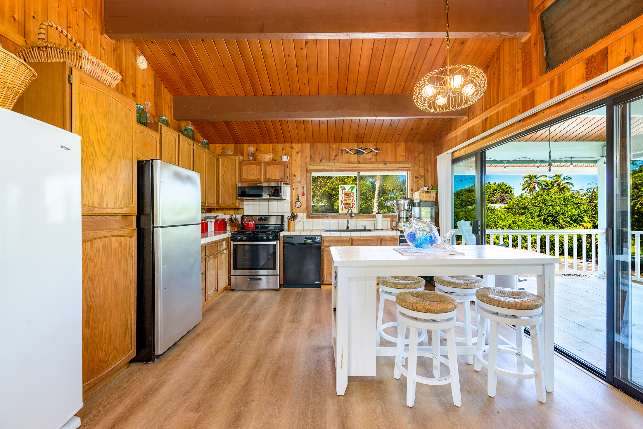 Cozy wooden kitchen with stainless appliances, white island, and stools. Glass doors open to a sunny patio with trees outside.