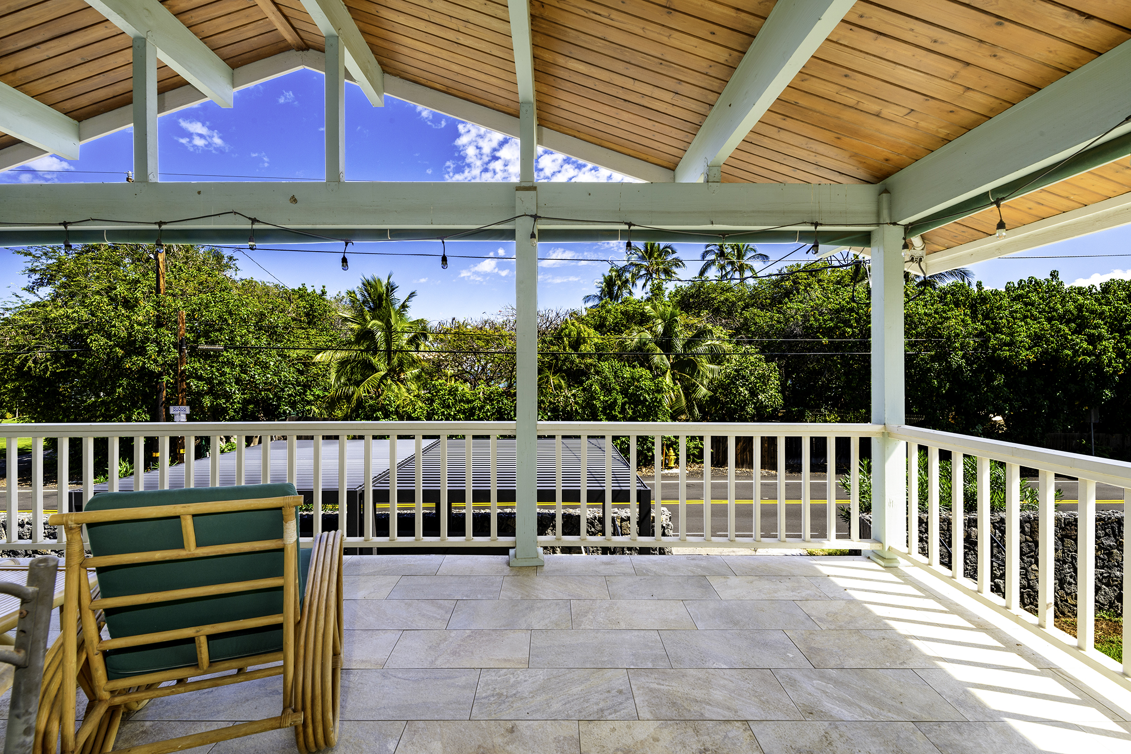 Covered patio with wooden ceiling, white railing, and a single wicker chair. Tropical greenery and clear blue sky in the background.