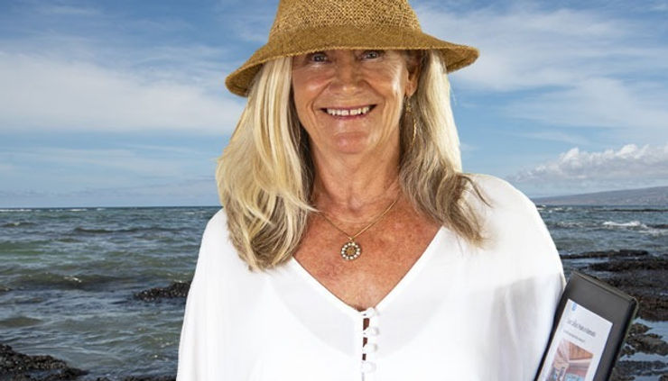 Woman in a white shirt with a brown hat smiling by the seaside. She's holding a tablet. The sky is clear, and the ocean is calm.