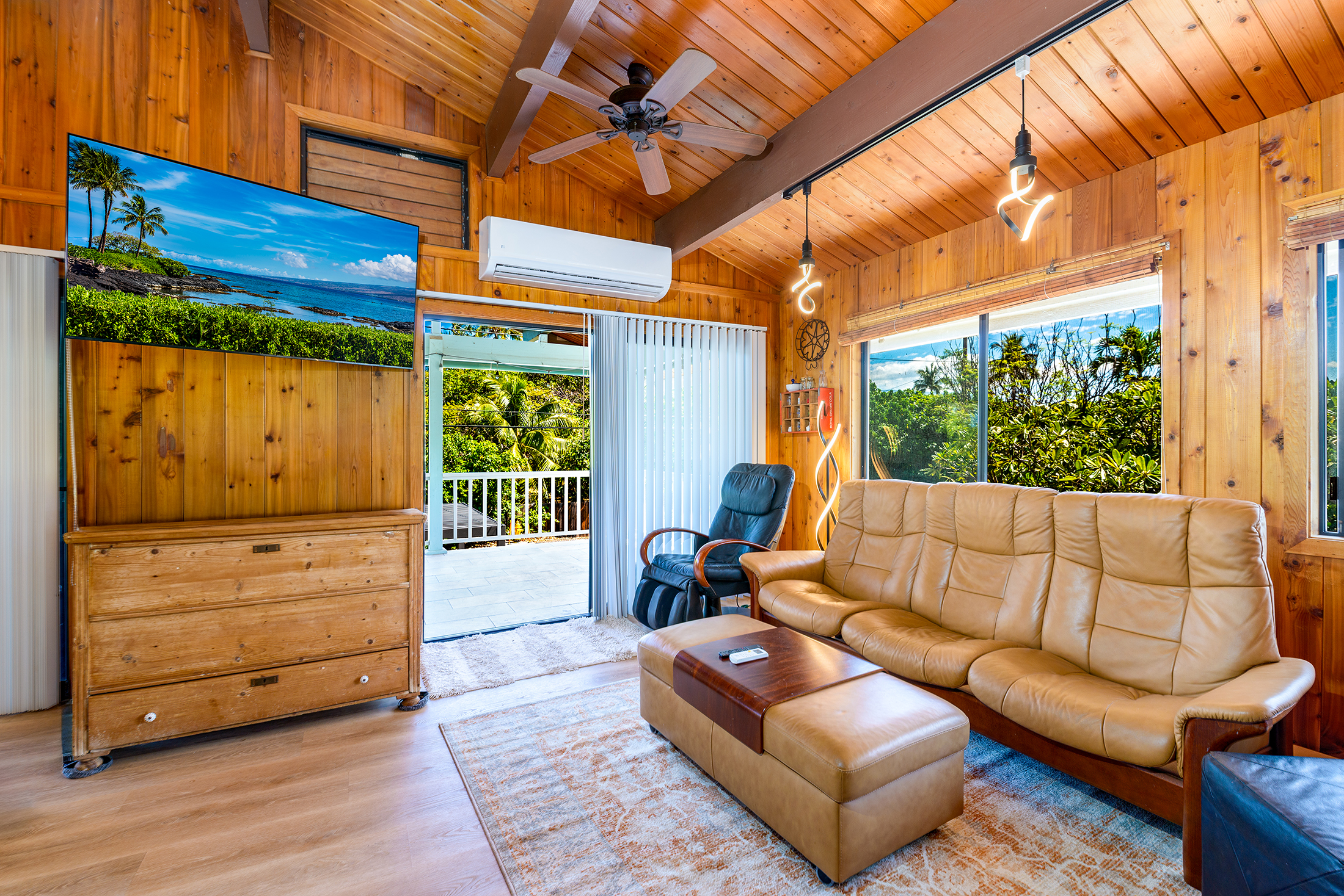 Wood-paneled living room with beige sofas, a TV showing a beach scene, large windows, and a view of lush greenery outside. Cozy atmosphere.