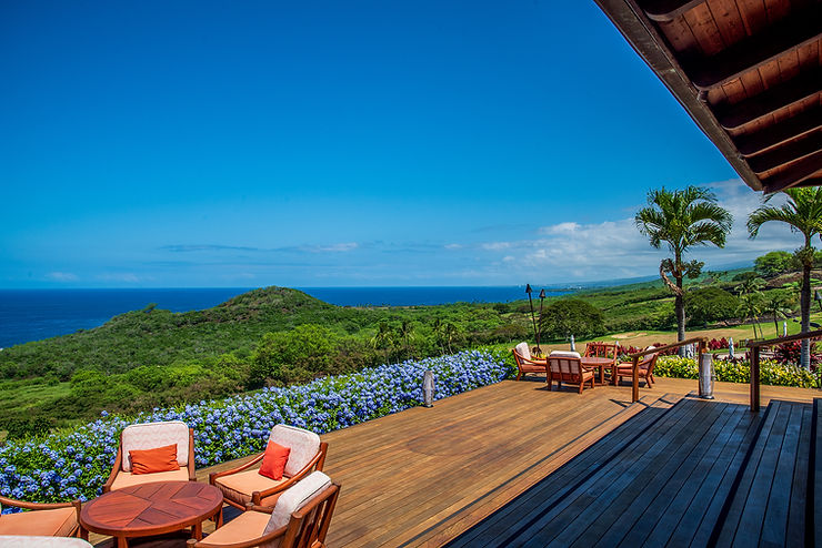 Ocean view from a wooden deck at Hokulia Club with chairs, tables, and vibrant flowers. Green hills and a bright blue sky in the background create a serene scene.