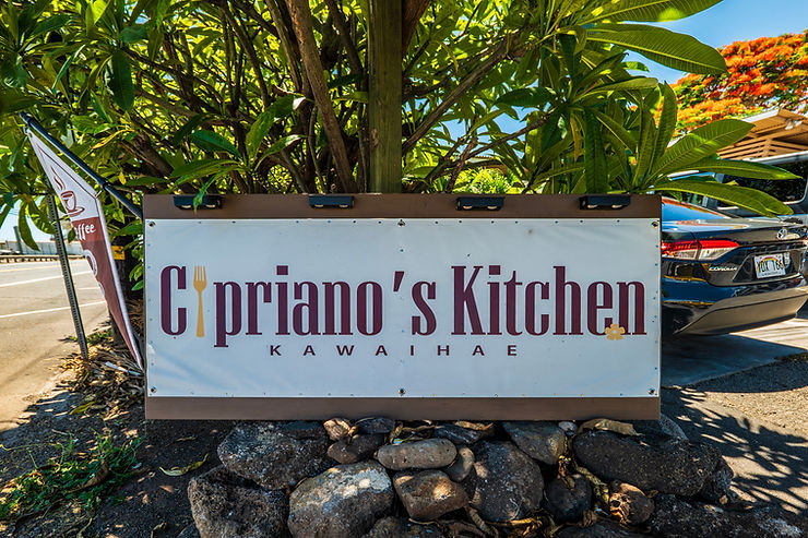 Capriano's Kitchen Kawaihae sign beside a parked car and tropical tree, with a coffee flag on a sunny street.