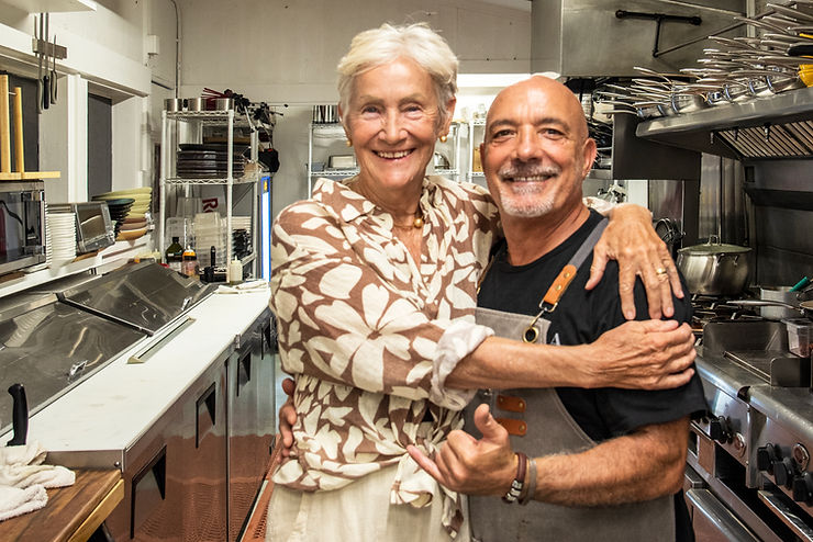 Smiling older woman hugs a chef in a stainless steel restaurant kitchen, both facing camera.