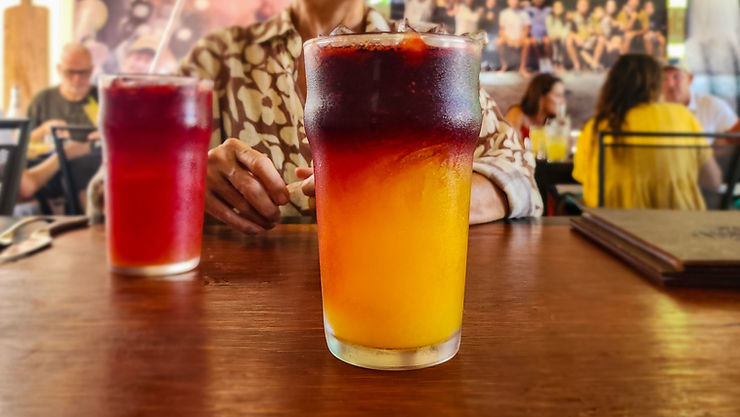 Two layered iced drinks on a wooden table in a busy restaurant, with blurred diners in the background.