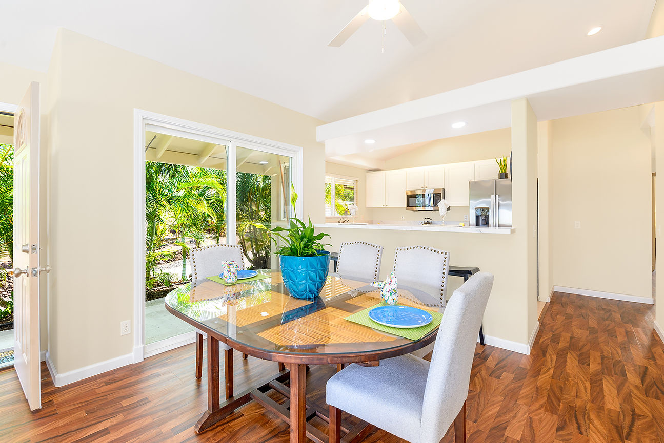 Bright dining area with glass table, gray chairs, blue planter, and open kitchen beside sliding doors to lush tropical greenery