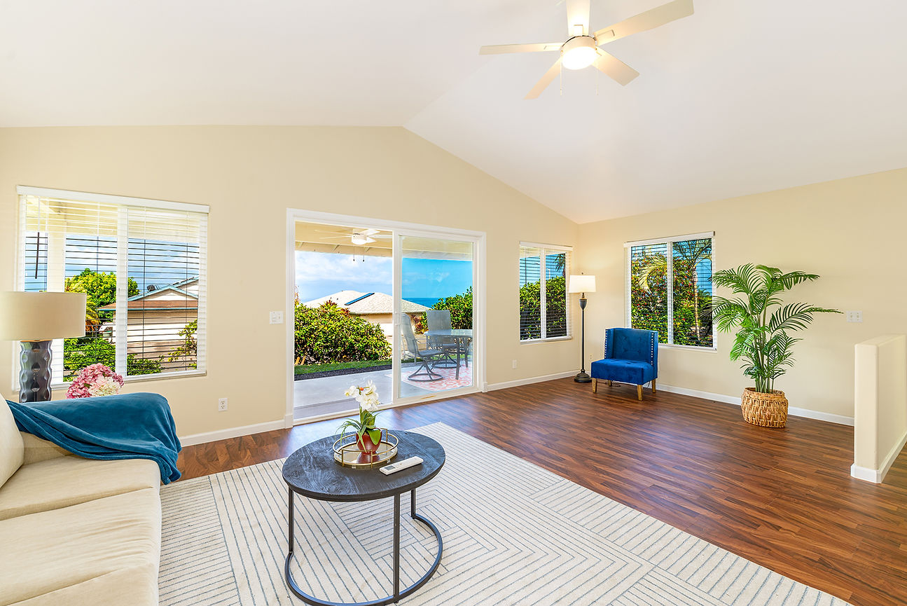 Bright living room with beige walls, blue chairs, sofa, plants and coffee table, opening to a sunny patio with ocean view.
