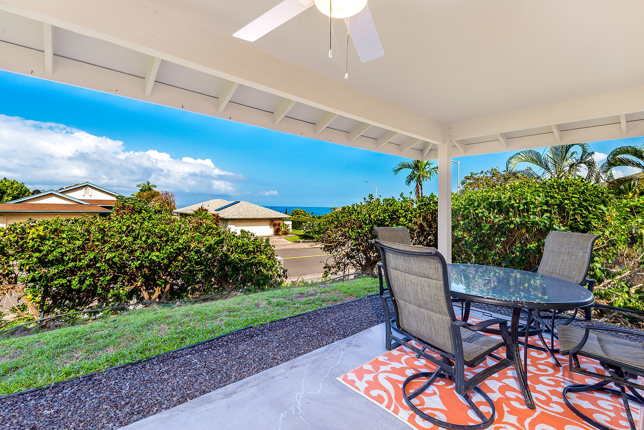 Covered patio with glass table and chairs overlooking tropical hedges, houses, and blue ocean under a bright sky
