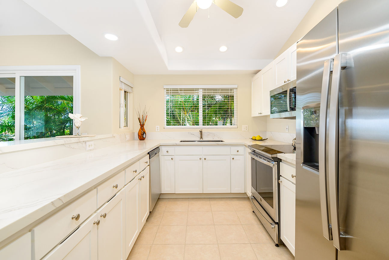 Bright modern white kitchen with stainless steel appliances, marble counters, and palm trees visible through the windows