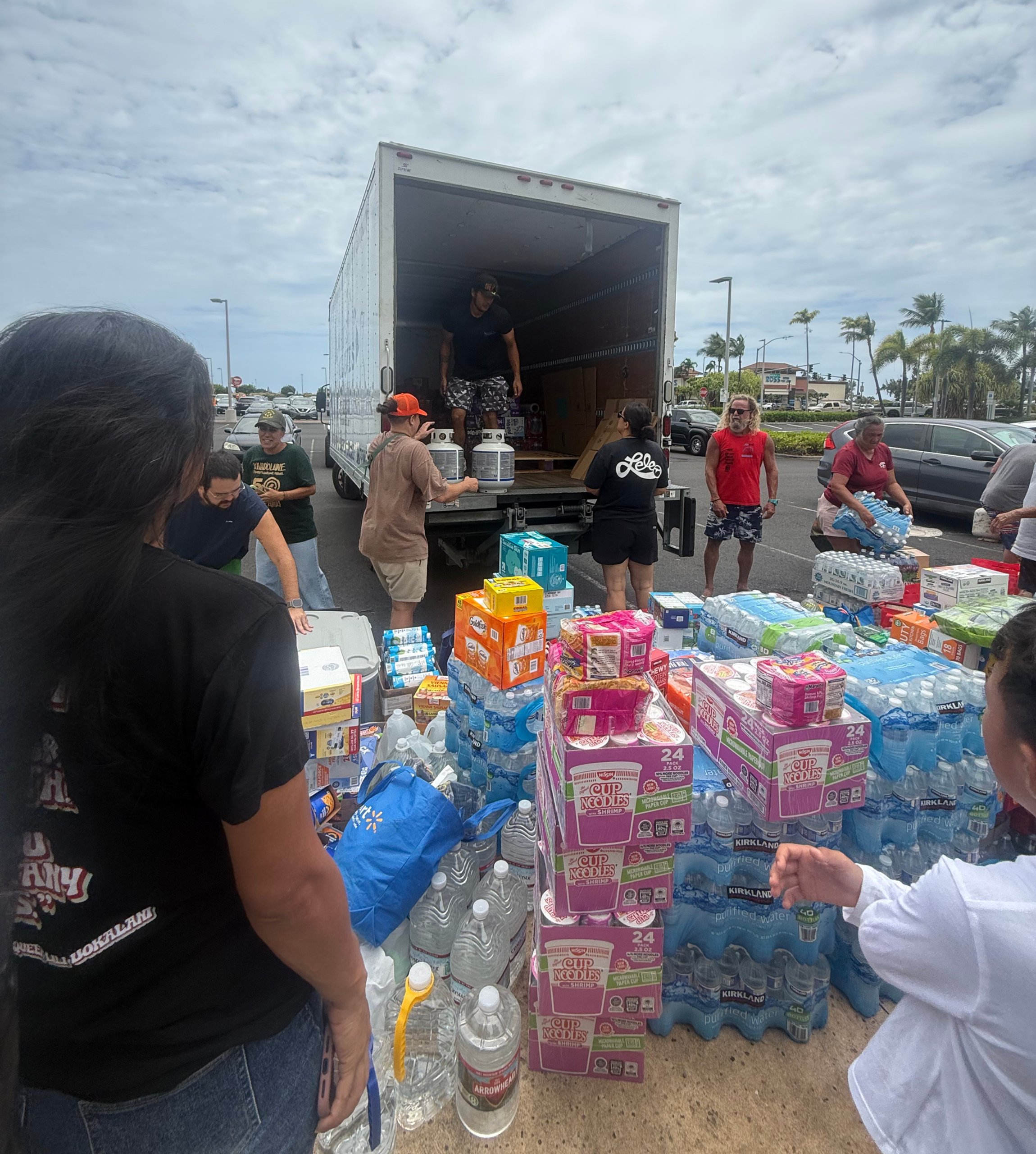 Volunteers unload water and instant noodles from a truck in a parking lot under cloudy skies, with stacked supplies and cars nearby.