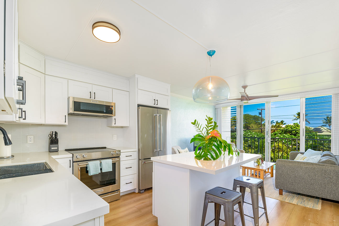 Bright modern kitchen with white cabinets, stainless appliances, island plant and stools, opening to a sunny living room and tropical view.
