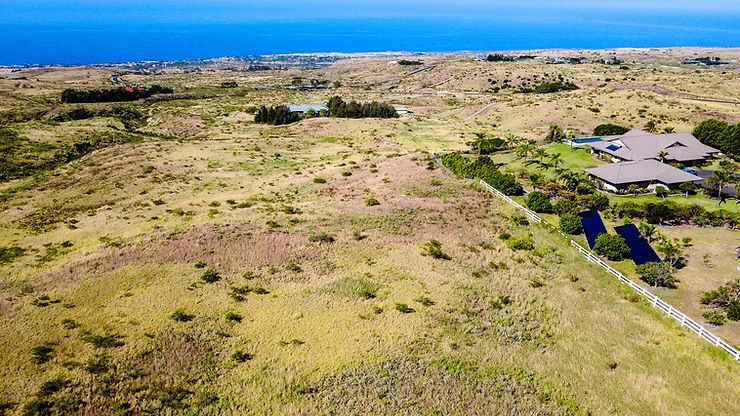 Aerial view of dry coastal hills and a large house with white fence beside blue ocean under clear sky.